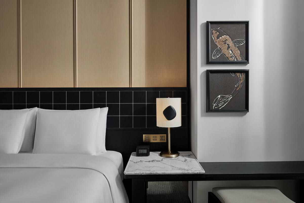 A hotel room with a bed, a nightstand with a lamp, a clock, and two framed artworks on the wall.