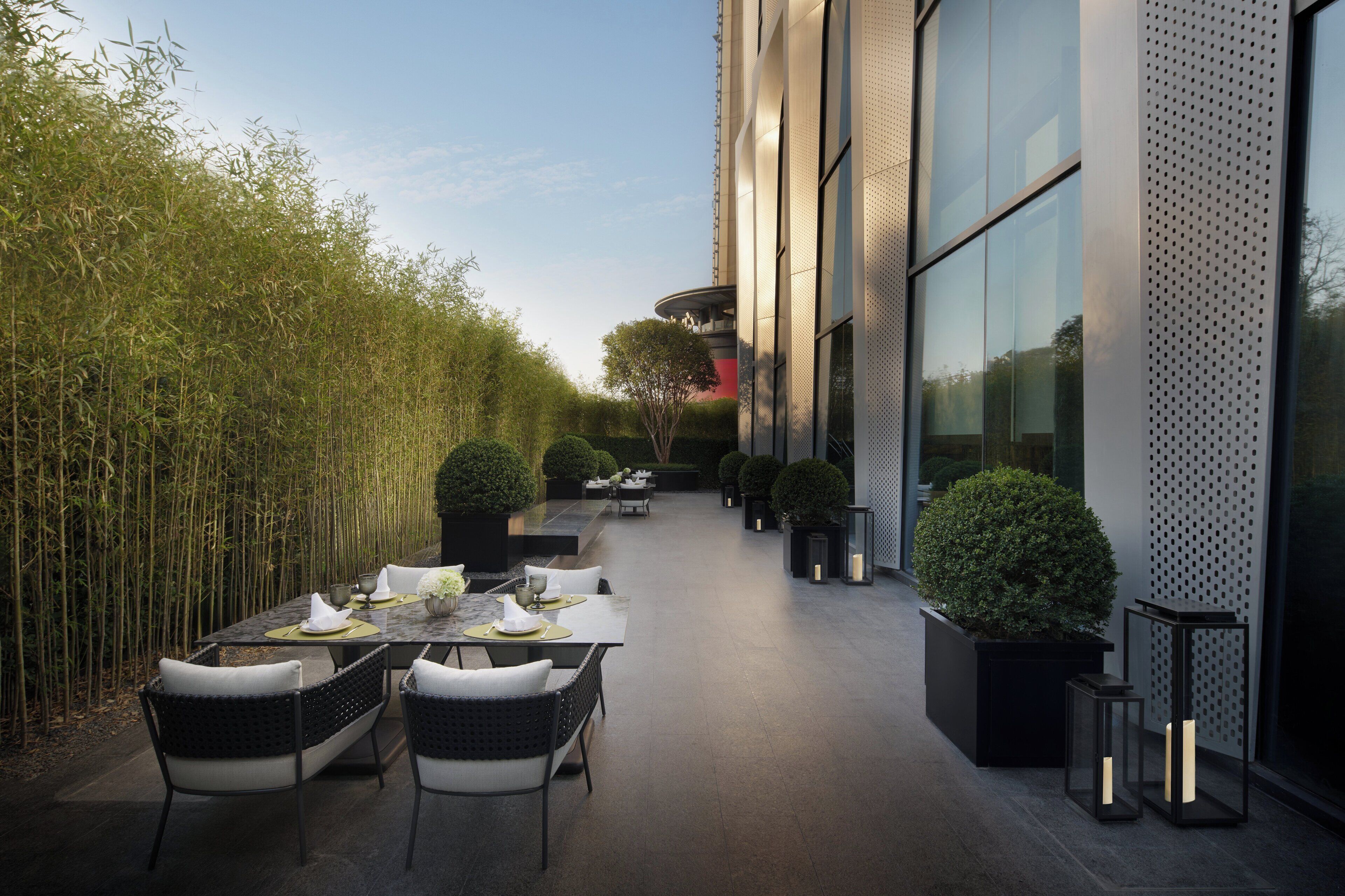 An outdoor dining area with tables and chairs, surrounded by bamboo plants and potted trees, leading to a modern building with large windows.