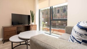 A modern living room with a flat-screen TV, a round table, a sofa, and a large window with a view of a brick building. - Liverpool Family Apartment 2 Bed/2 Bath (101) | Secure Parking, WiFi & EV (Liverpool)