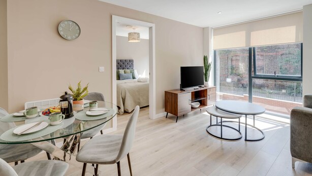 A modern living room with a glass dining table, a sofa, a TV on a wooden stand, and a large window offering a view of the outside. - Liverpool Family Apartment 2 Bed/2 Bath (101) | Secure Parking, WiFi & EV (Liverpool)