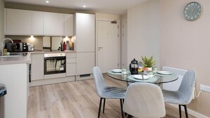 A modern kitchen with built-in appliances, a dining area with a glass table and chairs, and a wall clock. - Liverpool Family Apartment 2 Bed/2 Bath (101) | Secure Parking, WiFi & EV (Liverpool)