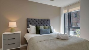 A bedroom with a tufted headboard, a bedside table with drawers, a lamp, and a large window with a view of a brick building. - Liverpool Family Apartment 2 Bed/2 Bath (101) | Secure Parking, WiFi & EV (Liverpool)