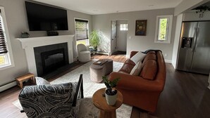 Een moderne woonkamer met een open haard, een grote flatscreen televisie, een bruine leren bank, een gestreept fauteuil en een kleine ronde tafel met een plant.
