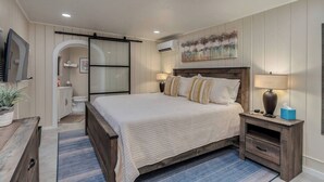 A bedroom with a large bed, a bedside table, a nightstand, a wall-mounted TV, and a bathroom with a toilet and sink. - Casita Sam Hughes/University King bed (Tucson)
