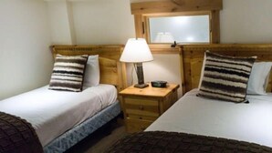 Ein Hotelzimmer mit zwei Einzelbetten, jeweils mit einem Holzkopfstück und gestreiften Kissen. Ein Nachttisch mit Lampe und ein kleiner Tisch befinden sich zwischen den Betten.