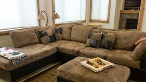 Ein Wohnzimmer mit einem Ecksofa, einem kleinen Hocker mit einer Snackplatte, einem gemusterten Teppich und einem Kamin im Hintergrund.