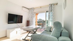 A living room with a flat-screen TV, a sofa, a dining table, and a balcony with a view of palm trees.
