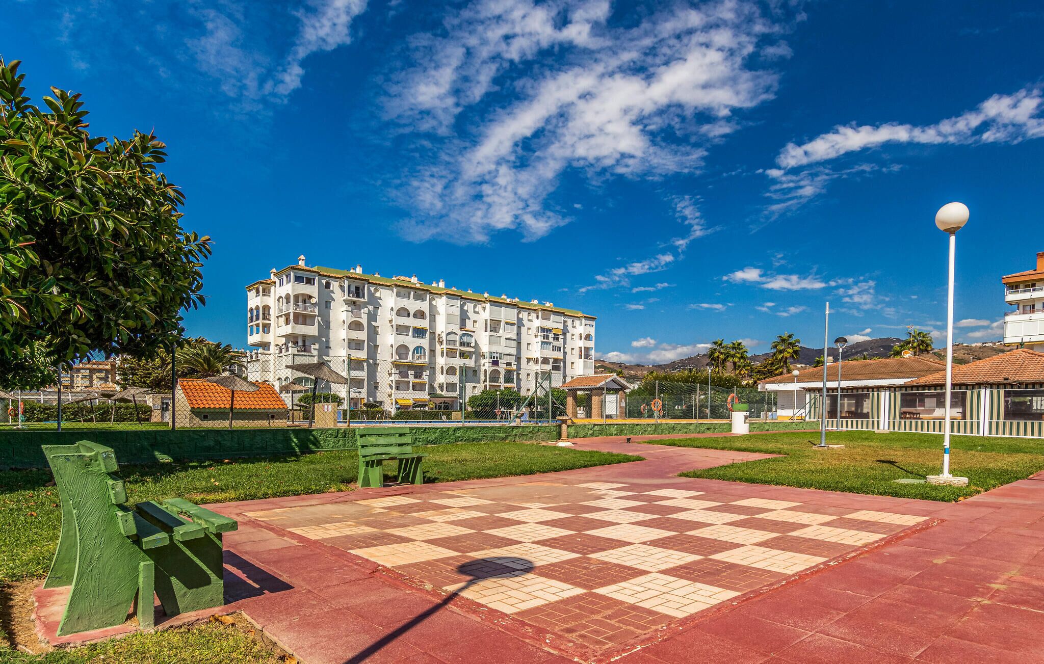 A park with a checkered pavement, green benches, and a building with multiple balconies in the background.