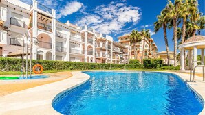 A swimming pool surrounded by multi-story buildings with balconies, palm trees, and a clear blue sky.
