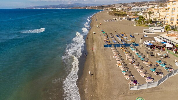 An aerial view of a beach with rows of sun loungers and umbrellas, a promenade, and buildings in the background.