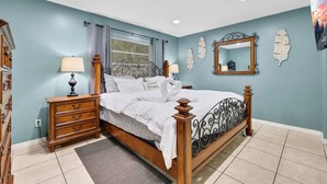 Une chambre à coucher comprenant un lit, des tables de chevet, une commode, une fenêtre avec des rideaux, un miroir et des éléments décoratifs sur les murs.