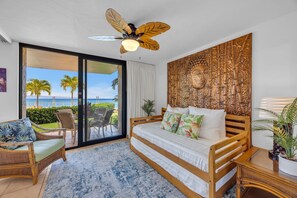 Une chambre avec un lit, un ventilateur de plafond, une vue sur l’océan et une décoration murale sur le thème des tropiques.