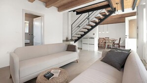 A modern living room with a white sofa, a round woven ottoman, and a staircase with a black railing. - Amazing home in Funtana (Funtana)