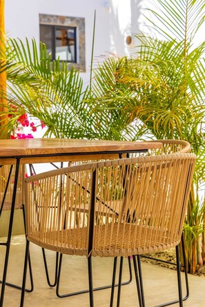Une table et des chaises en osier sont disposées à l’extérieur, avec un bâtiment blanc et de la verdure en arrière-plan.