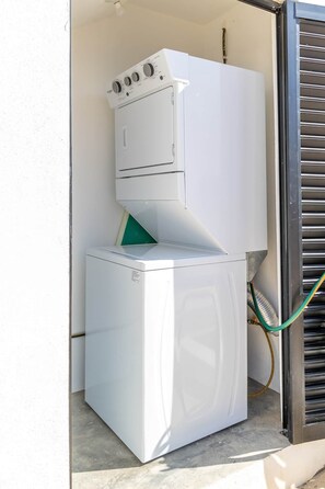 Une petite machine à laver blanche, dotée d’un tuyau, est placée contre un mur dans un espace extérieur étroit.