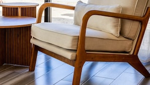 A modern chair with a cushion, a round wooden table, and a view of a desert landscape through a large window. - El Corazón Contento | Cerritos Beach (La Paz)