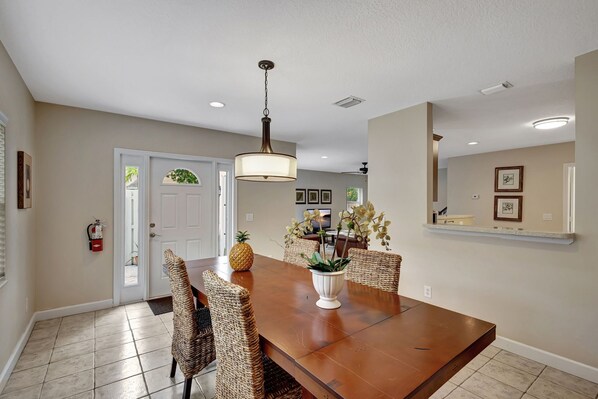 A dining area with a wooden table, wicker chairs, a white vase with flowers, and a hanging light fixture.