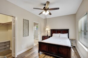 Un dormitorio con cama de madera, ventilador de techo, lámpara de noche y ventana con persianas.
