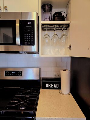A kitchen with a stainless steel microwave, a stove, and a shelf with wine glasses and a Hamilton Beach blender.
