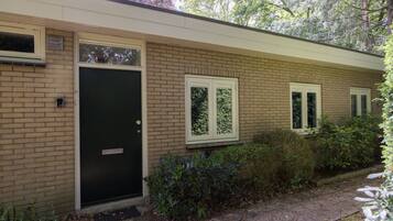 Een bakstenen huis met een zwarte deur, twee ramen met witte kaders, en een kleine tuin.