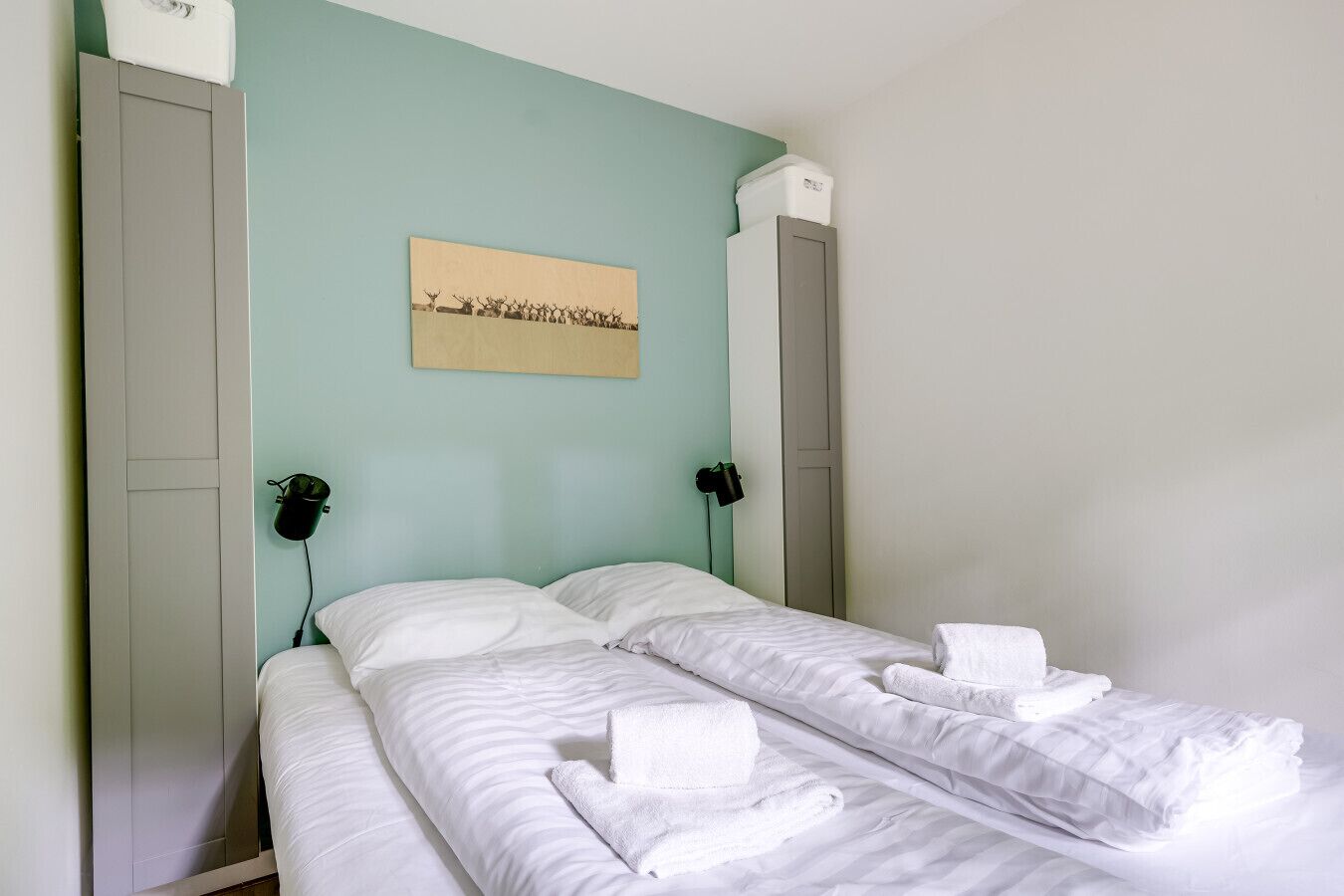 Un lit simple avec du linge de lit et des serviettes blanches, un mur vert clair et un tableau encadré au-dessus du lit.