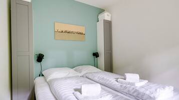 Un lit simple avec du linge de lit et des serviettes blanches, un mur vert clair et un tableau encadré au-dessus du lit.