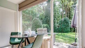 Een kamer met een tafel en stoelen, een raam met uitzicht op een tuin, en een deur die naar buiten leidt.