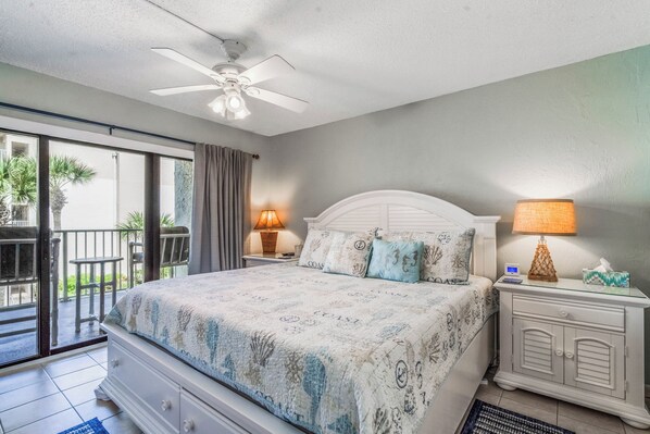 A bedroom with a bed, two nightstands, a ceiling fan, a balcony with a chair, and a view of the outdoors. - Castle Reef 327 beautiful, lush, views! (New Smyrna Beach)