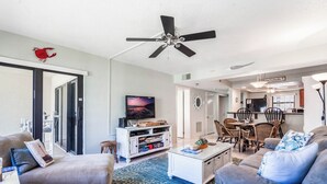 Ein Wohnzimmer mit Deckenventilator, Fernseher, Essbereich mit Stühlen und einer Küche im Hintergrund.