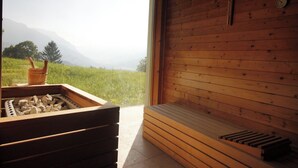 A sauna room with a wooden bench and a stone basin, a wooden wall, and a view of a green landscape with mountains in the distance.