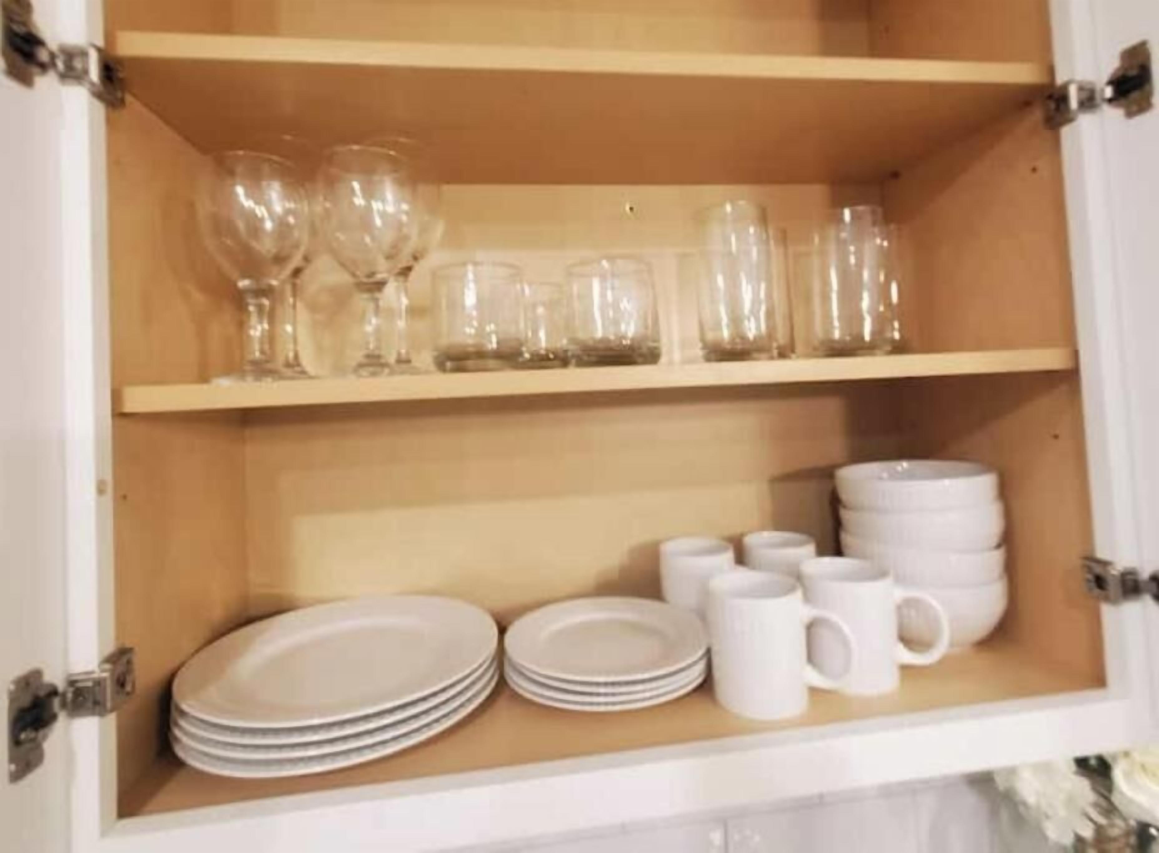 A cabinet with two shelves; the top shelf holds clear glassware, and the bottom shelf contains stacked white plates and white mugs.