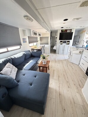 Interior compacto de una caravana con sofá, una mesita con un florero, una zona de cocina con armarios y un televisor sobre la encimera.