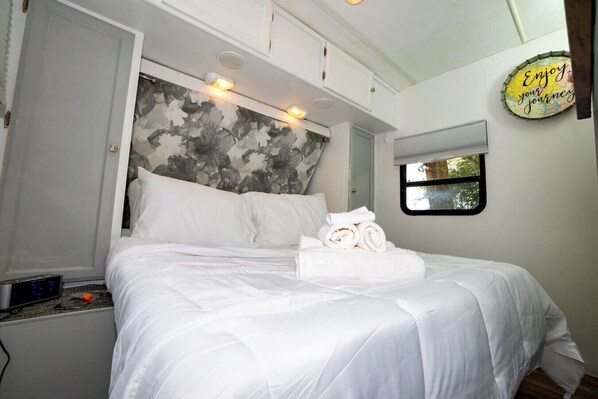 A neatly made bed with white linens and rolled towels, a digital clock, and a wall clock on the bedside table.