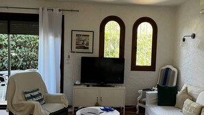 A living room with a television, a sofa, a chair, and a small table. There are two windows with arched tops and a door leading outside. - Villa T4 type canétoise air-conditioned and near beach - 6ILEF56 (CANET PLAGE)