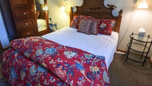 Een slaapkamer met een houten hoofdbord, een bed met bloemenbeddengoed, een nachtkastje met een lamp en een houten garderobe.