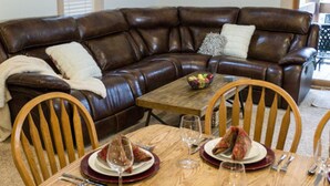 A brown leather sectional sofa with a white throw blanket and pillows, a wooden dining table with plates and wine glasses, and two wooden chairs. - 4BR Spacious Condo | Private Deck & Fireplace (Truckee)