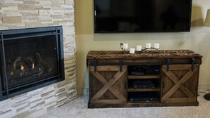 A living room with a stone fireplace, a flat-screen TV mounted on the wall, and a wooden entertainment center with a candle holder. - 4BR Spacious Condo | Private Deck & Fireplace (Truckee)