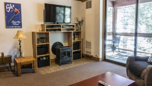 A living room with a television, wooden shelves, a fireplace, and a large window with a view of trees. - Spacious Studio Loft | Fireplace + Private Balcony (Truckee)