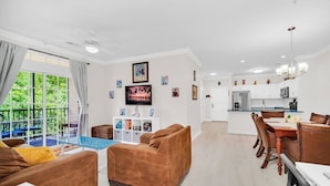 A modern living room with a sofa, a coffee table, and a TV. There is a dining area with a table and chairs. The room has a large window with curtains.