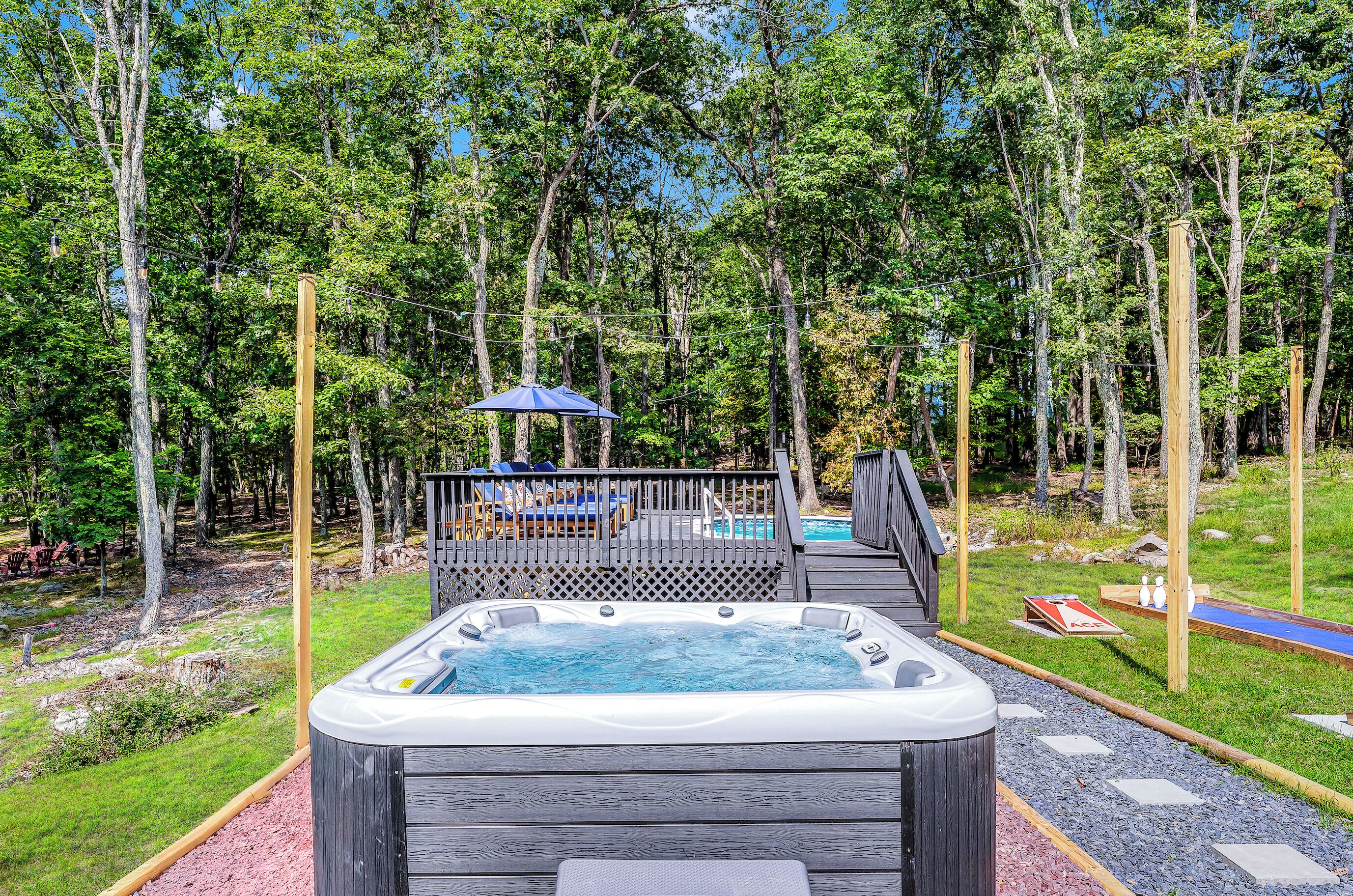 Een hottub op een houten terras, met een zwembad en ligbedden in een bebost gebied.