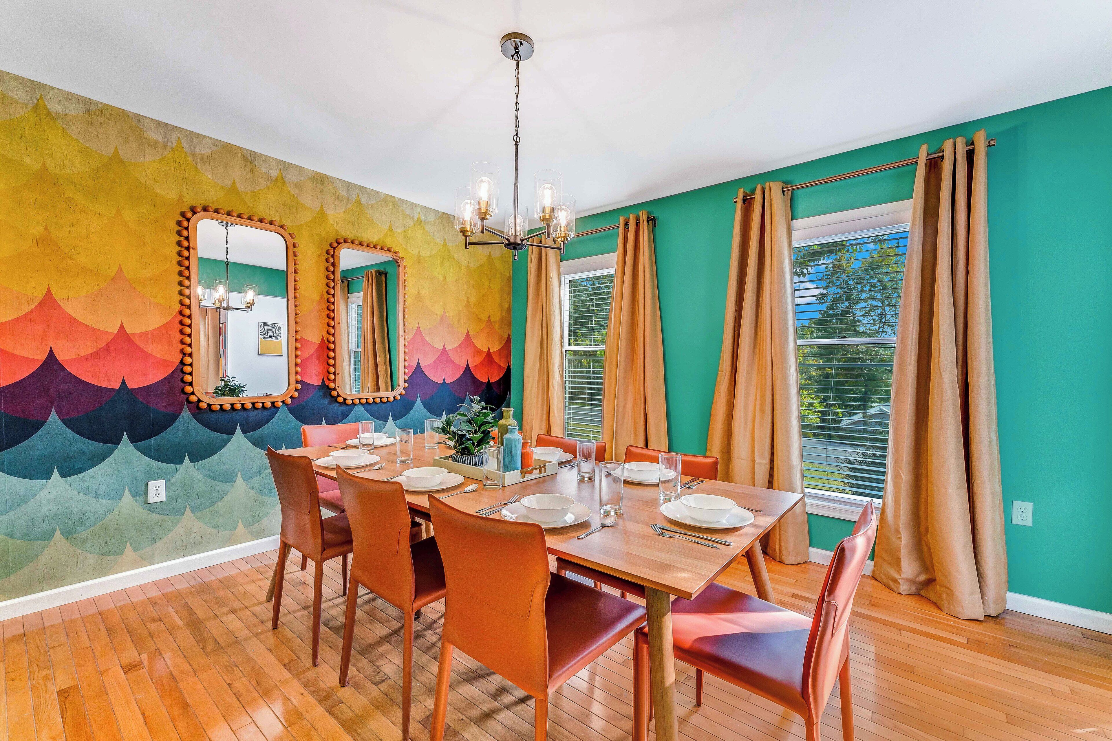 Un comedor con una mesa de madera dispuesta para cuatro personas, rodeada de sillas naranjas. Un mural colorido decora la pared detrás de la mesa.