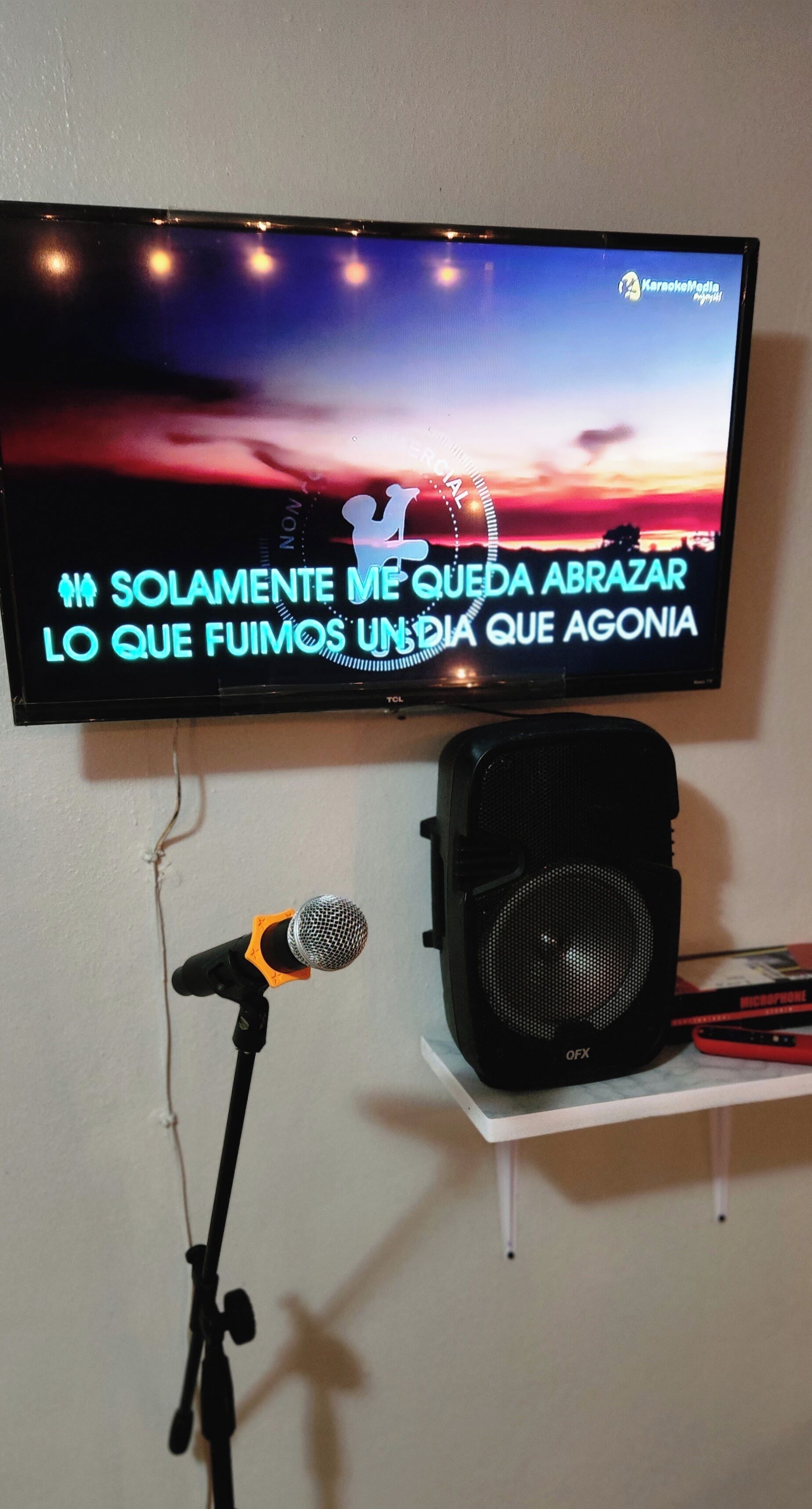A karaoke setup with a television displaying a song, a microphone, and a speaker on a shelf.
