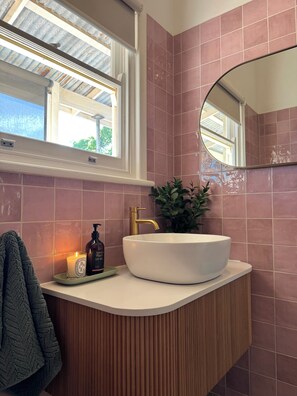 A bathroom with a white sink, a gold faucet, a mirror, a window, and a potted plant.
