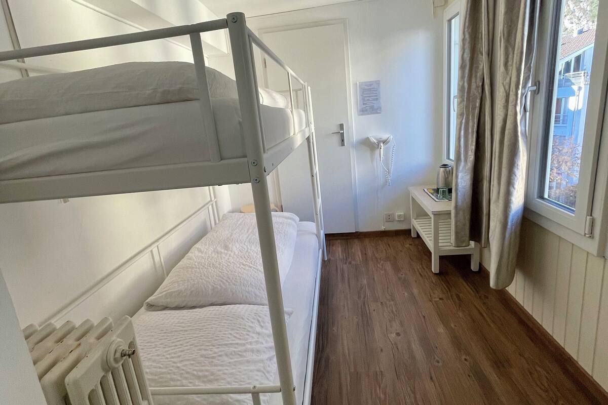 A bunk bed room with a wooden floor, a small bedside table, a radiator, and a window with curtains.