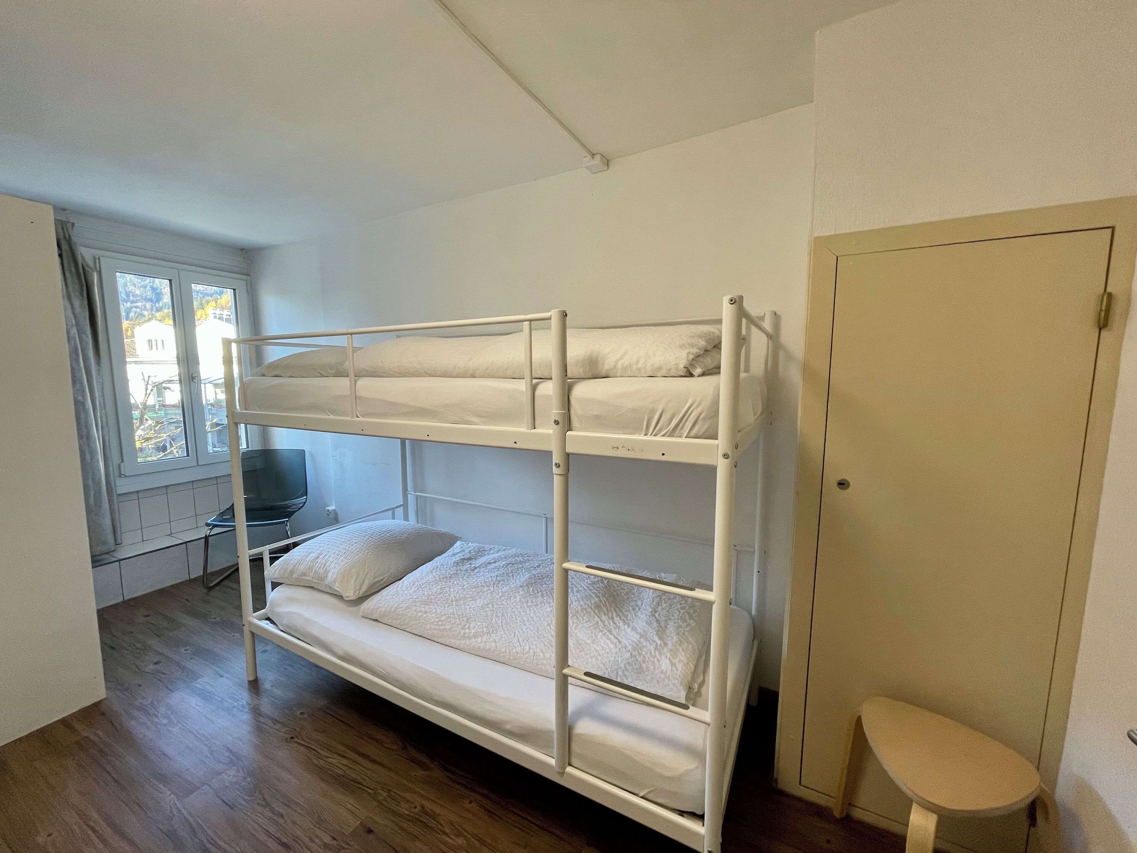 A bunk bed room with a desk and chair, a window with curtains, and a door.
