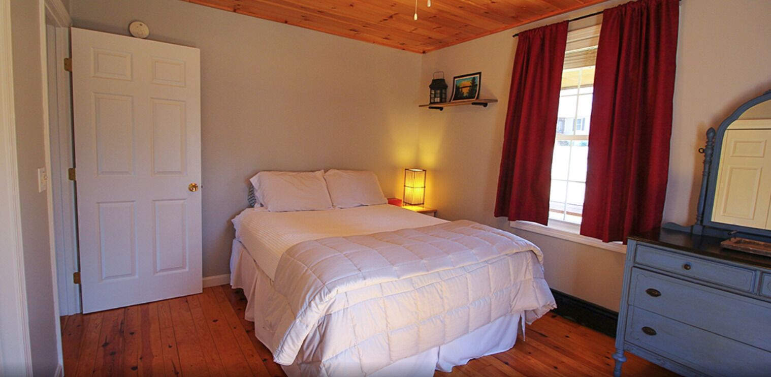 Une chambre à coucher comprenant un lit, une commode, une table de chevet avec une lampe, une porte et une fenêtre avec des rideaux.