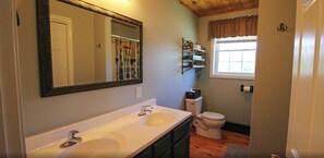 A bathroom with a double sink, a toilet, a mirror, and a window with a curtain. - Rangeley Getaway (Rangeley)