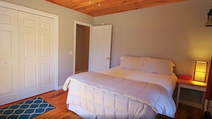 A bedroom with a bed, a nightstand, a lamp, and a door leading to another room. - Rangeley Getaway (Rangeley)