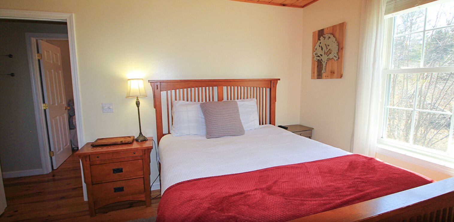 A bedroom with a wooden bed, a nightstand, a lamp, and a window with a view of trees.