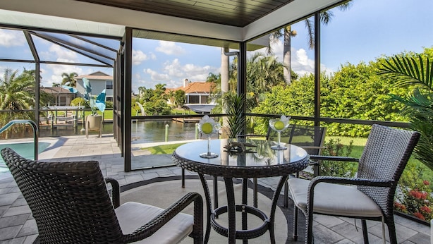 A covered outdoor seating area with wicker chairs and a glass table, overlooking a swimming pool and lush greenery.